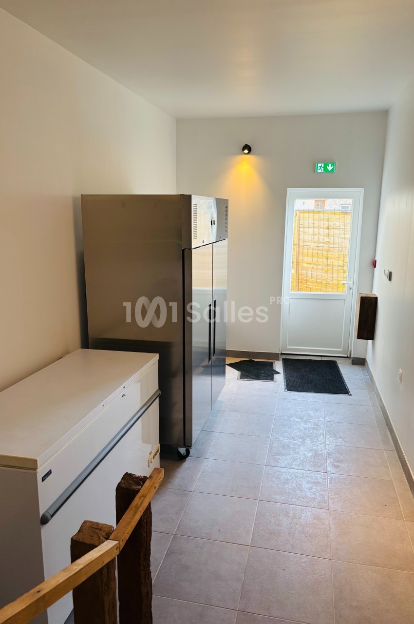 Couloir lumineux avec un congélateur, un réfrigérateur inox et une porte vitrée donnant sur l'extérieur.