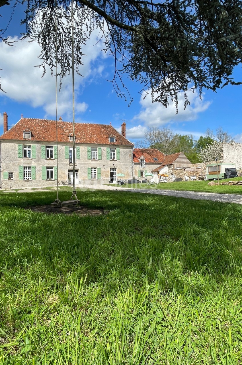 Grande maison ancienne en pierre avec toit en tuiles rouges, entourée d'un jardin verdoyant sous un ciel partiellement…