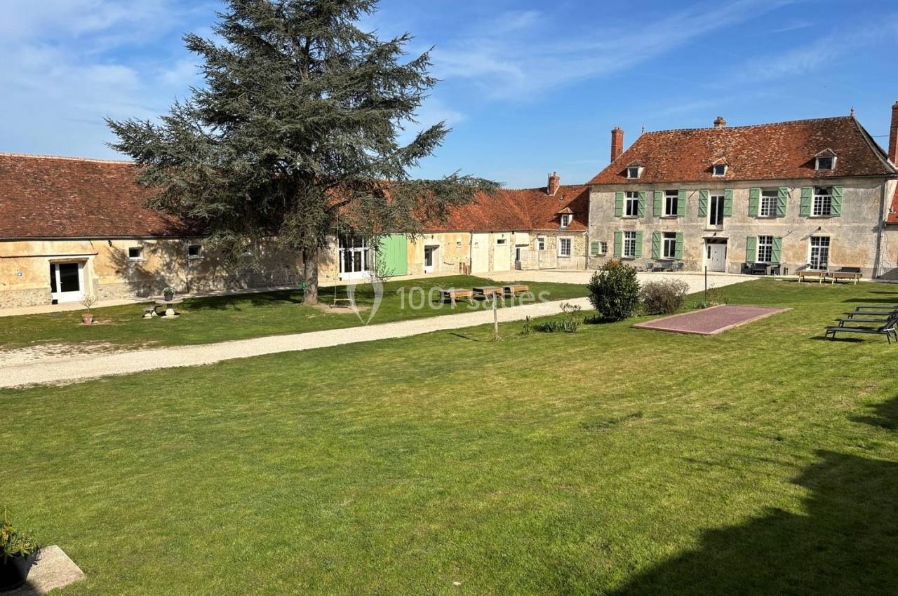 Cour verdoyante avec pelouse, bancs, arbre central et bâtiments anciens en pierre sous un ciel dégagé.