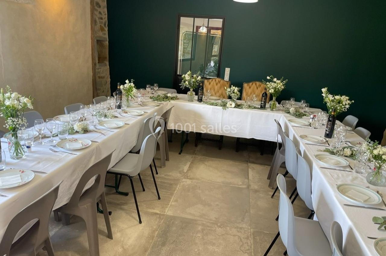 Salle décorée pour un repas, avec des tables en U dressées de nappes blanches, vaisselle et bouquets de fleurs.