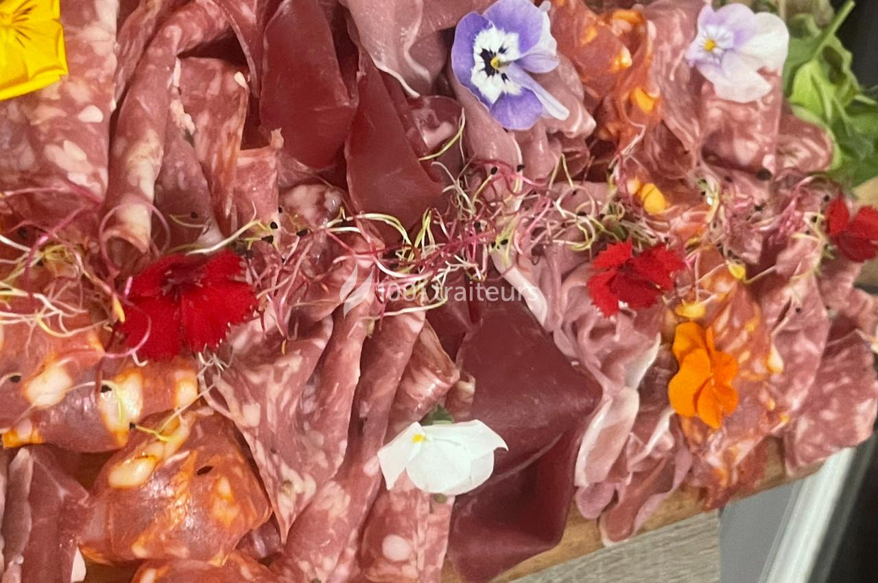 Plateau de charcuterie variée décoré de fleurs comestibles colorées et de pousses, présenté sur une planche en bois.