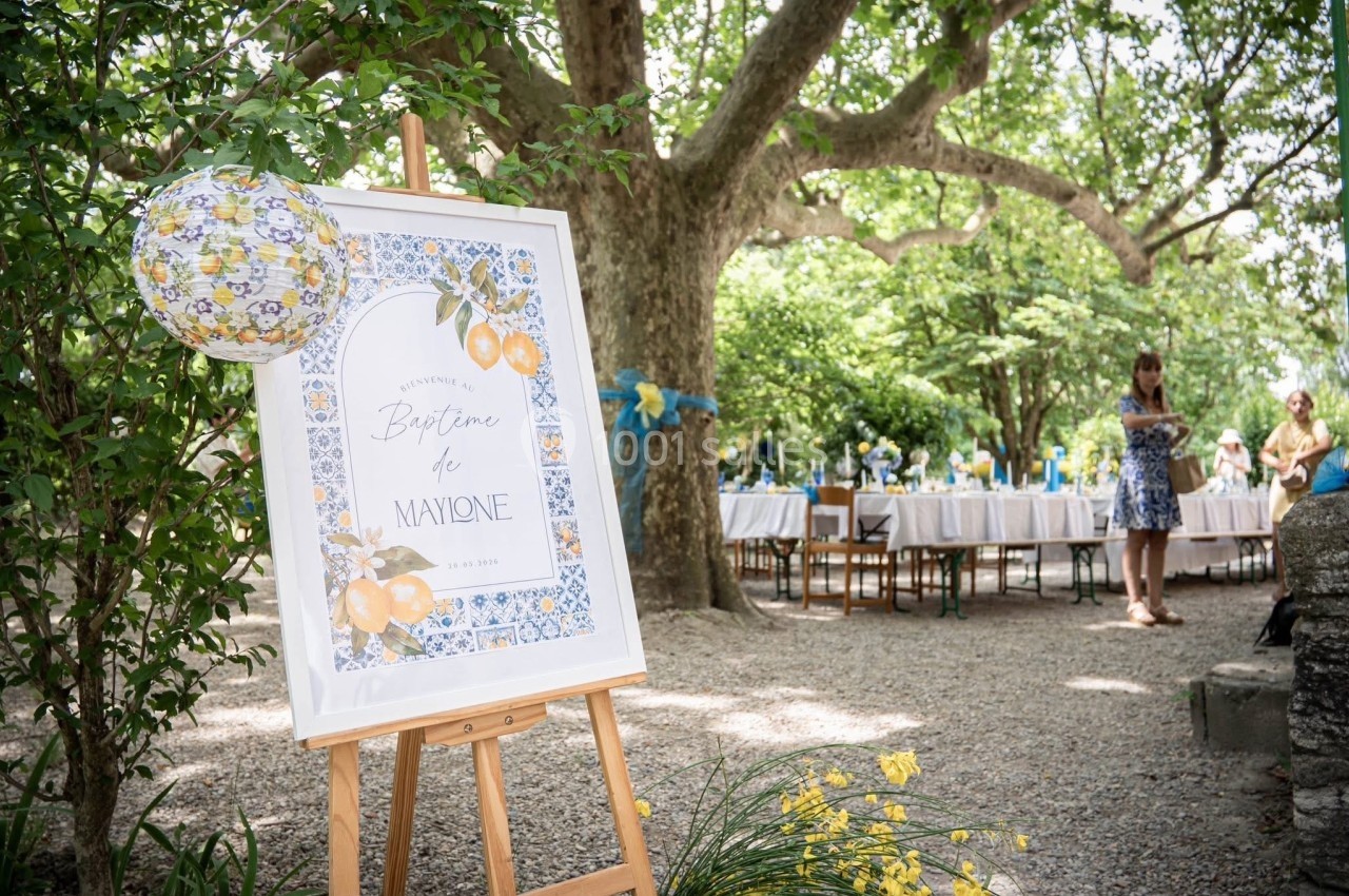 Panneau décoratif annonçant un baptême, posé sur un chevalet dans un jardin ombragé avec des tables dressées en arrière-plan.