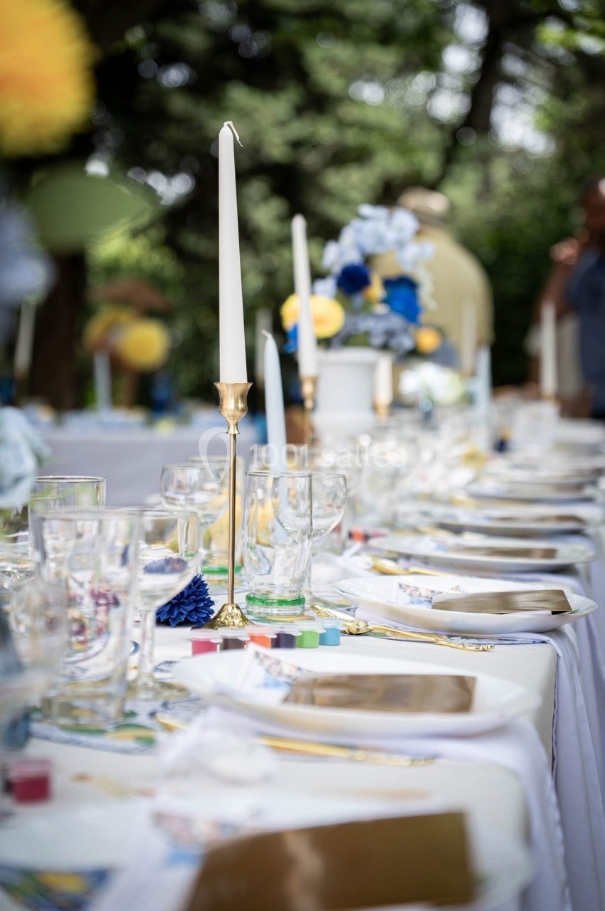 Table dressée en extérieur avec nappes blanches, bougies, verres et décorations florales bleues et jaunes.