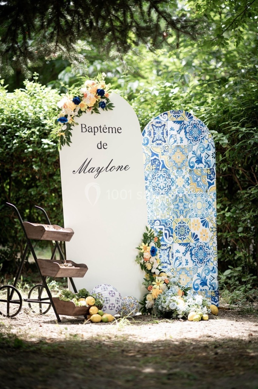 Décor de baptême avec panneau personnalisé, motifs colorés, fleurs, citrons et une étagère en bois dans un jardin.