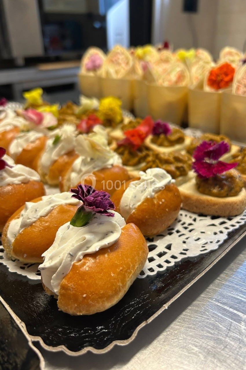 Plateau de petits fours variés, décorés de fleurs comestibles, disposés sur une nappe en dentelle blanche.