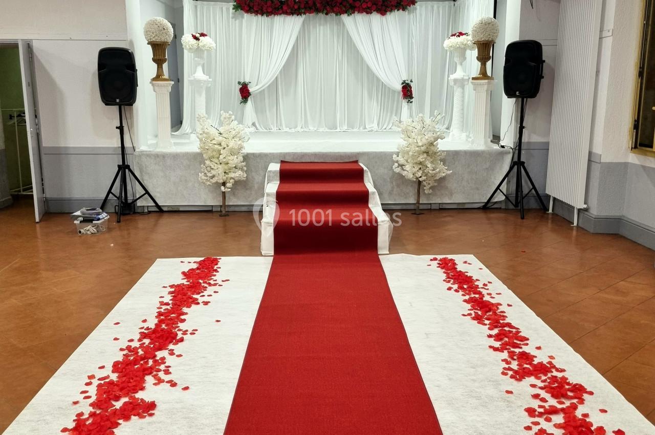 Allée rouge bordée de pétales de roses menant à une scène décorée de fleurs blanches et rouges dans une salle de réception.