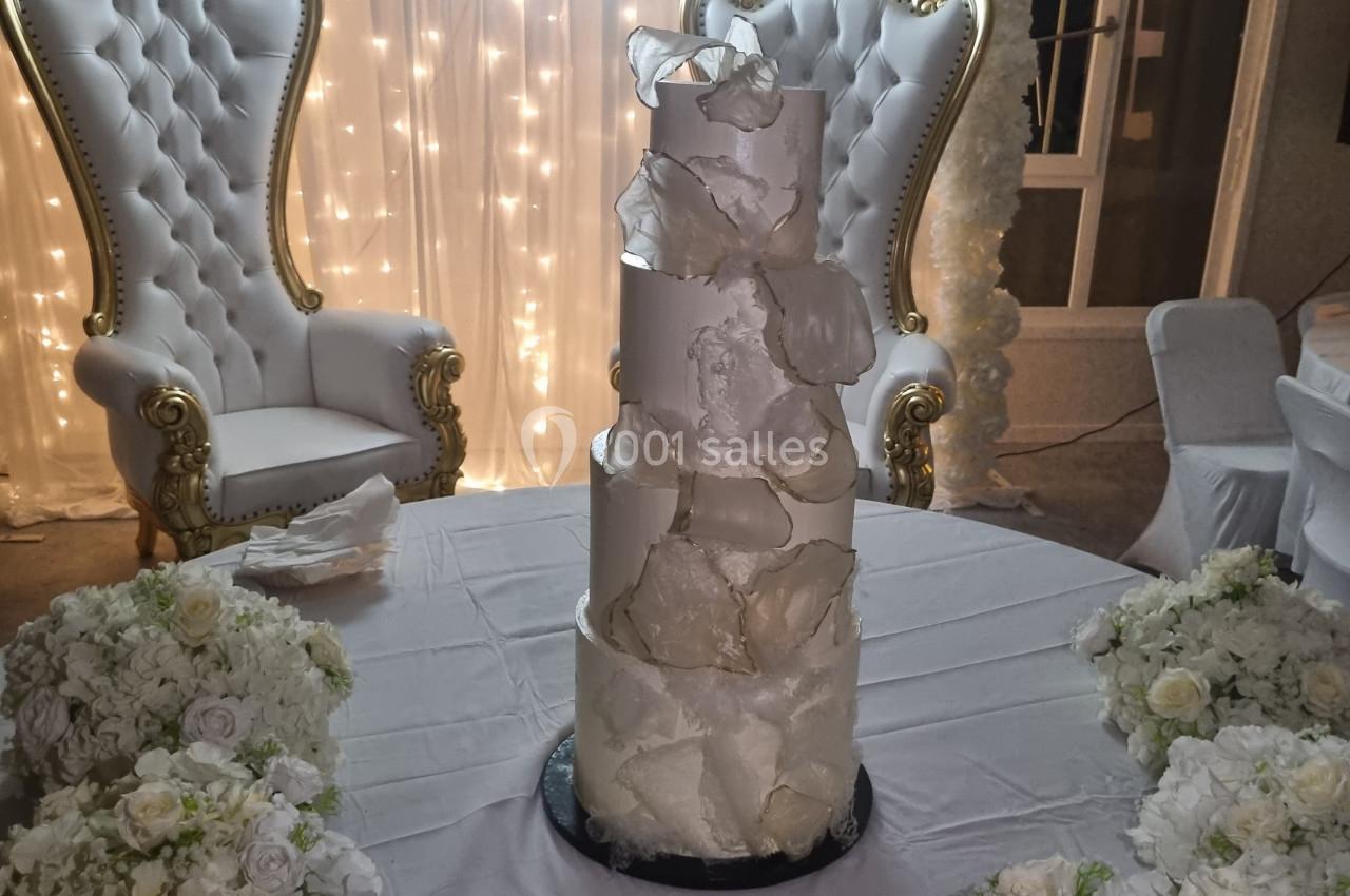 Gâteau de mariage à plusieurs étages décoré de blanc, entouré de bouquets de fleurs, avec deux fauteuils ornés en arrière…