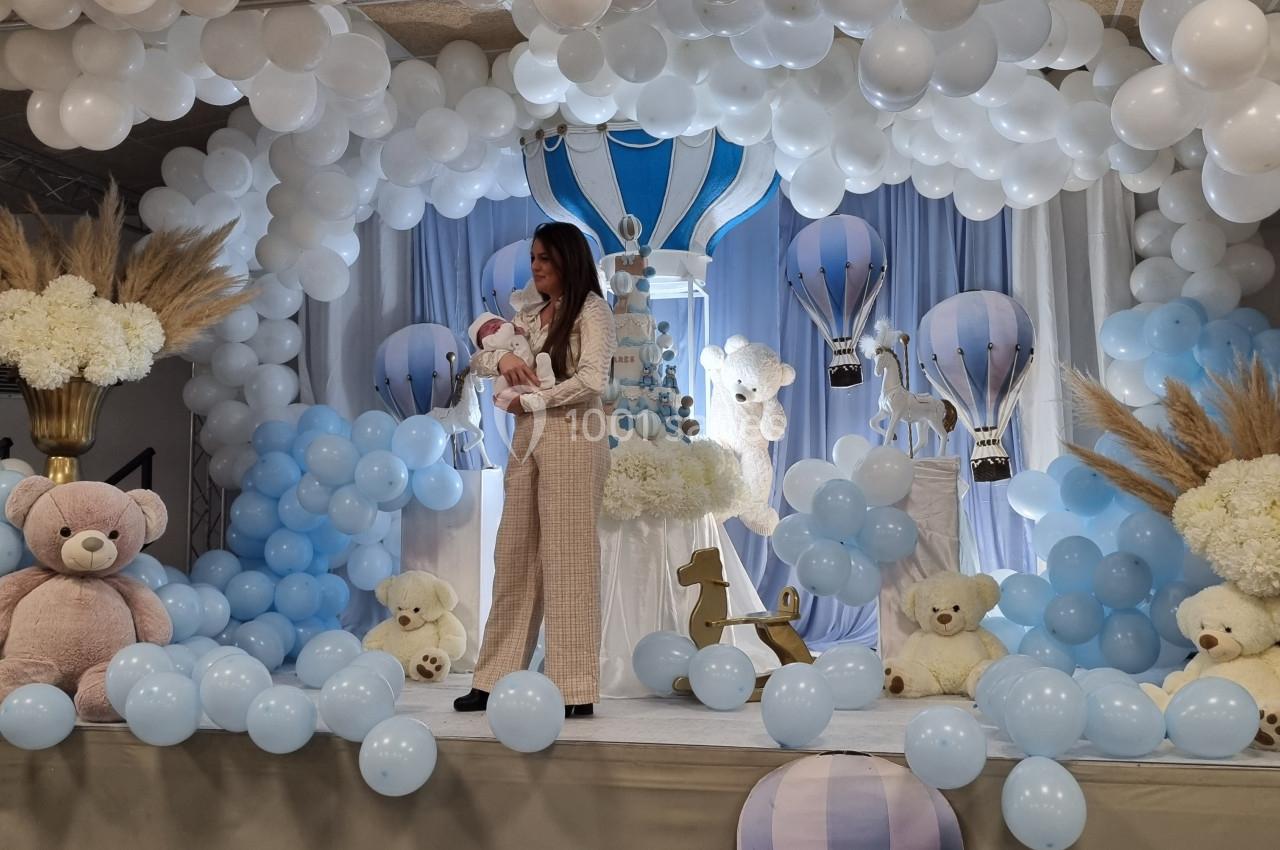 Une femme tient un bébé sur une scène décorée de ballons blancs et bleus, avec des peluches et des montgolfières.