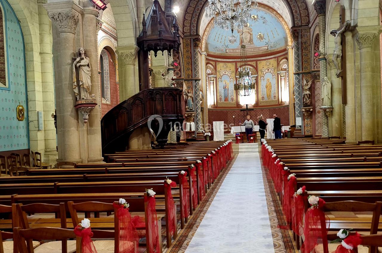 Intérieur d'une église décorée pour une cérémonie, avec des bancs ornés de rubans rouges et un autel en arrière-plan.