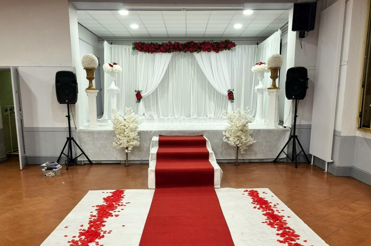 Scène décorée pour un mariage avec rideaux blancs, fleurs rouges et blanches, et tapis rouge parsemé de pétales.