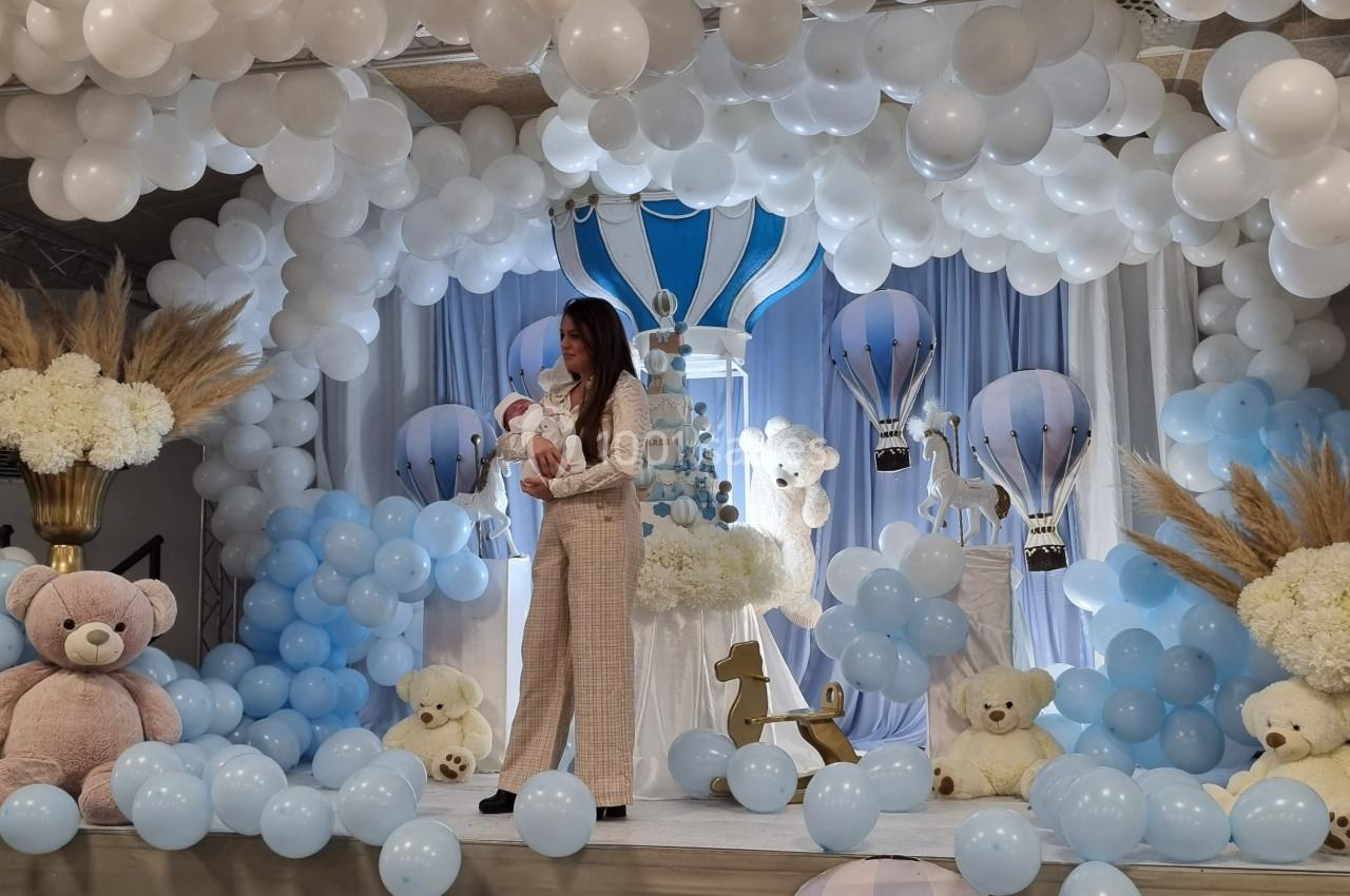 Une femme tenant un bébé dans une salle décorée de ballons bleus et blancs, avec des peluches et des montgolfières.
