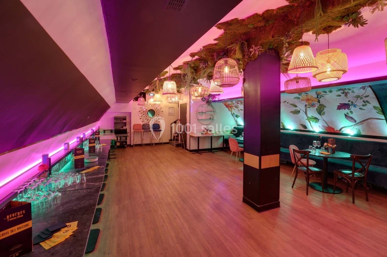 Salle lumineuse avec décoration végétale suspendue, tables et chaises colorées, et un bar sur la gauche.
