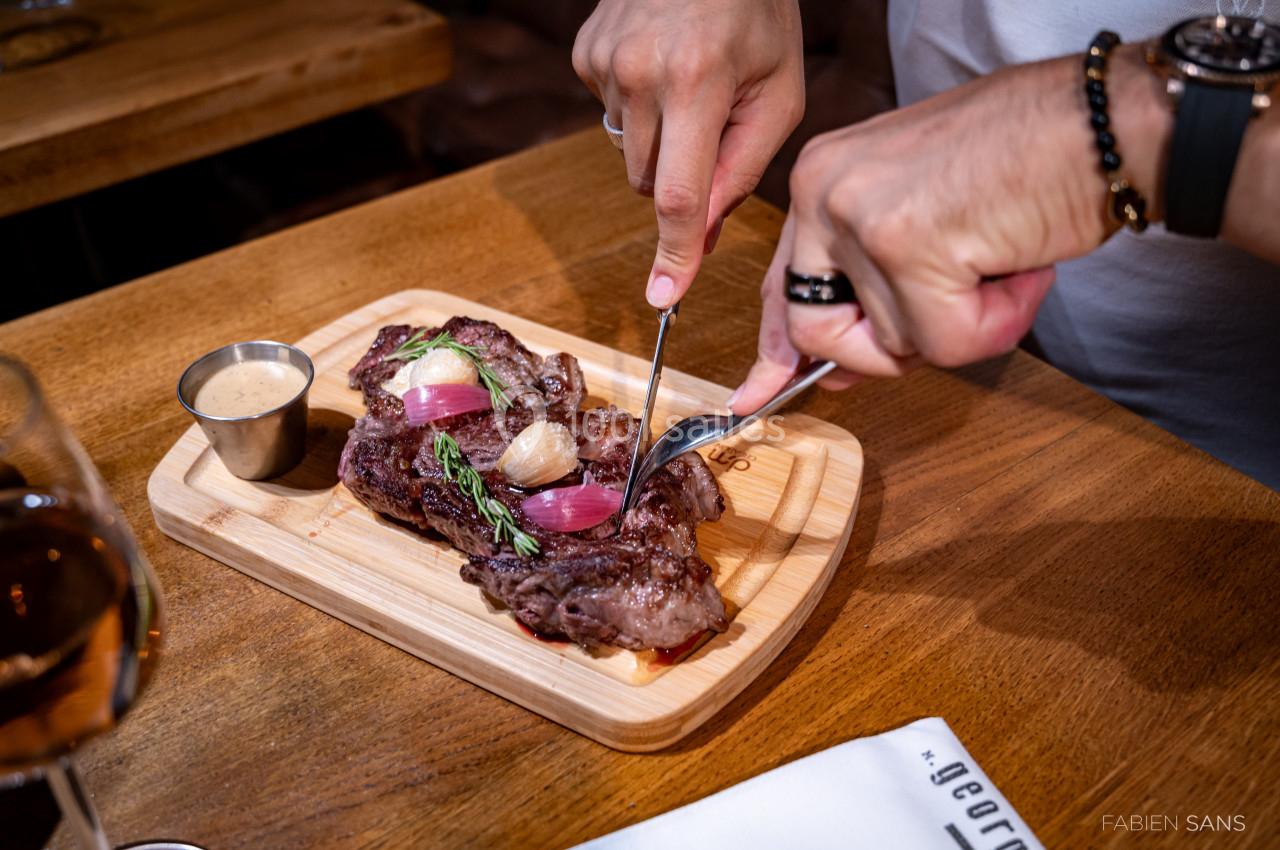 Une personne coupe une pièce de viande grillée garnie d'oignons et d'herbes, servie sur une planche en bois avec une sauce.