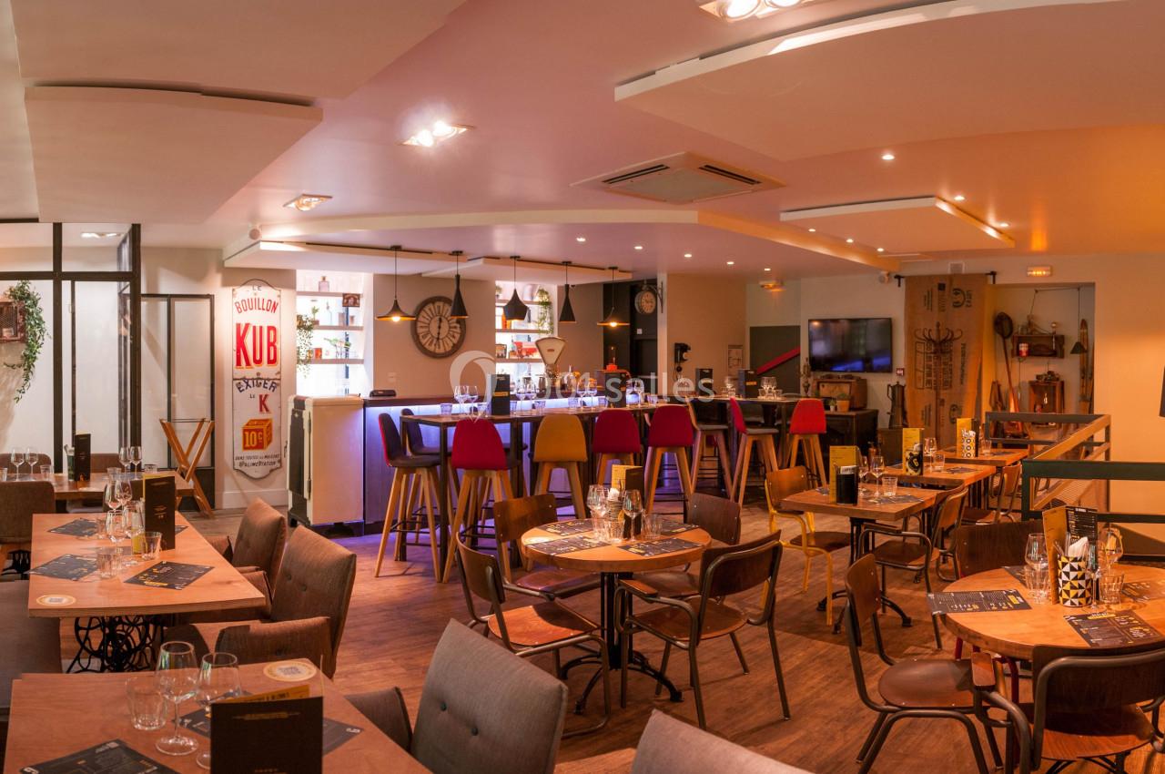 Salle de restaurant lumineuse avec tables en bois, bar coloré et décoration murale métallique.