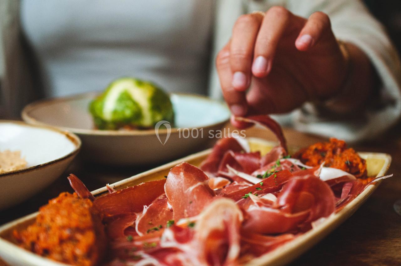 Assiette de charcuterie avec tranches de jambon cru et accompagnements, une main en train de se servir.