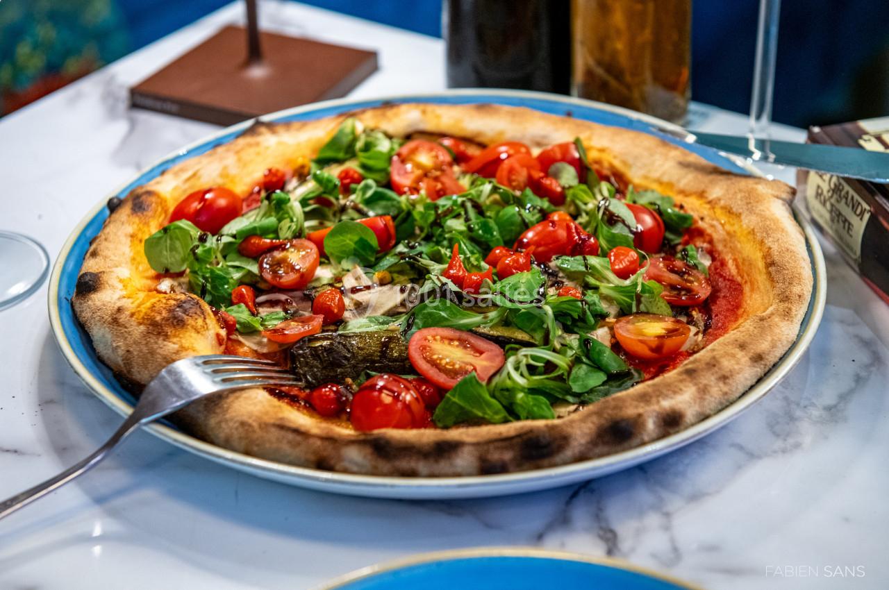 Pizza garnie de roquette, tomates cerises et légumes grillés, servie dans une assiette sur une table en marbre.