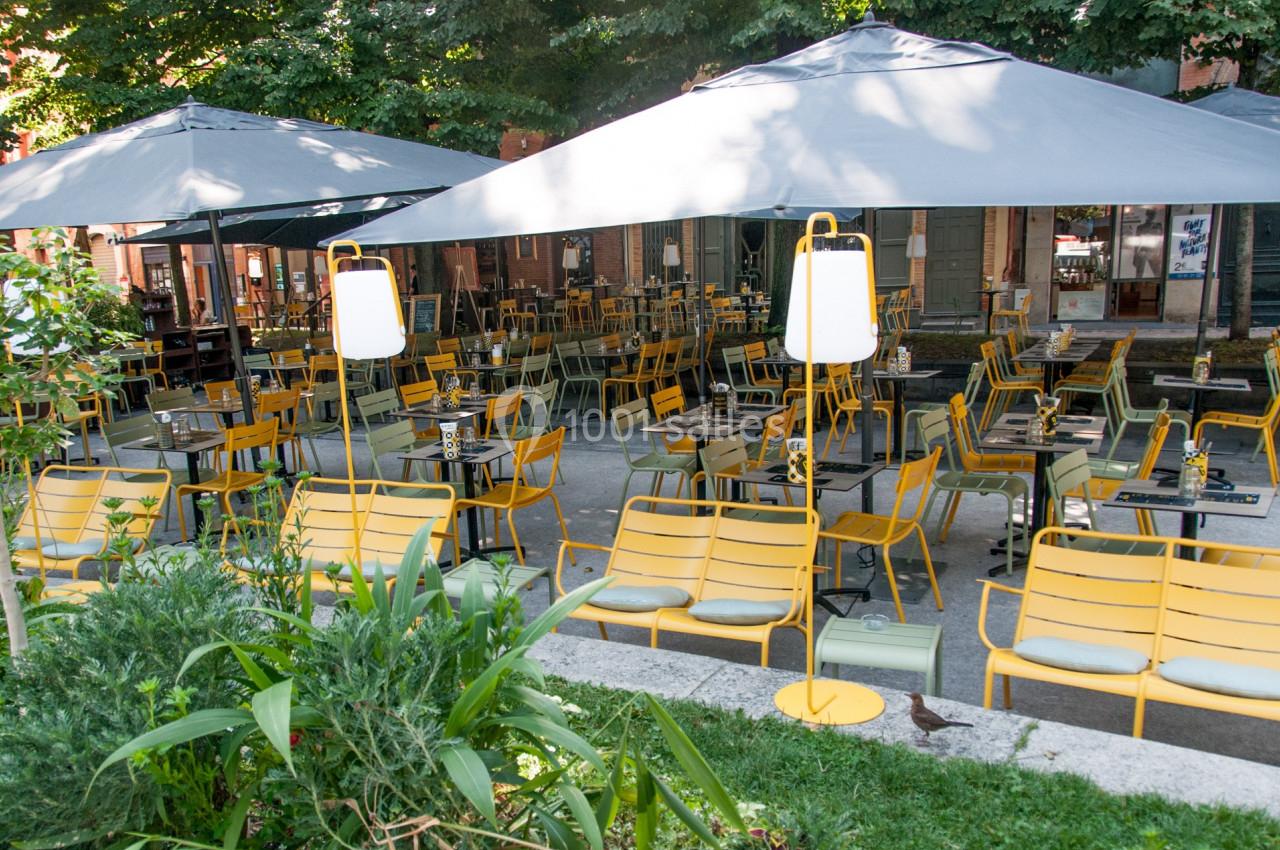 Terrasse extérieure avec des tables et chaises jaunes sous de grands parasols gris, entourée de verdure.
