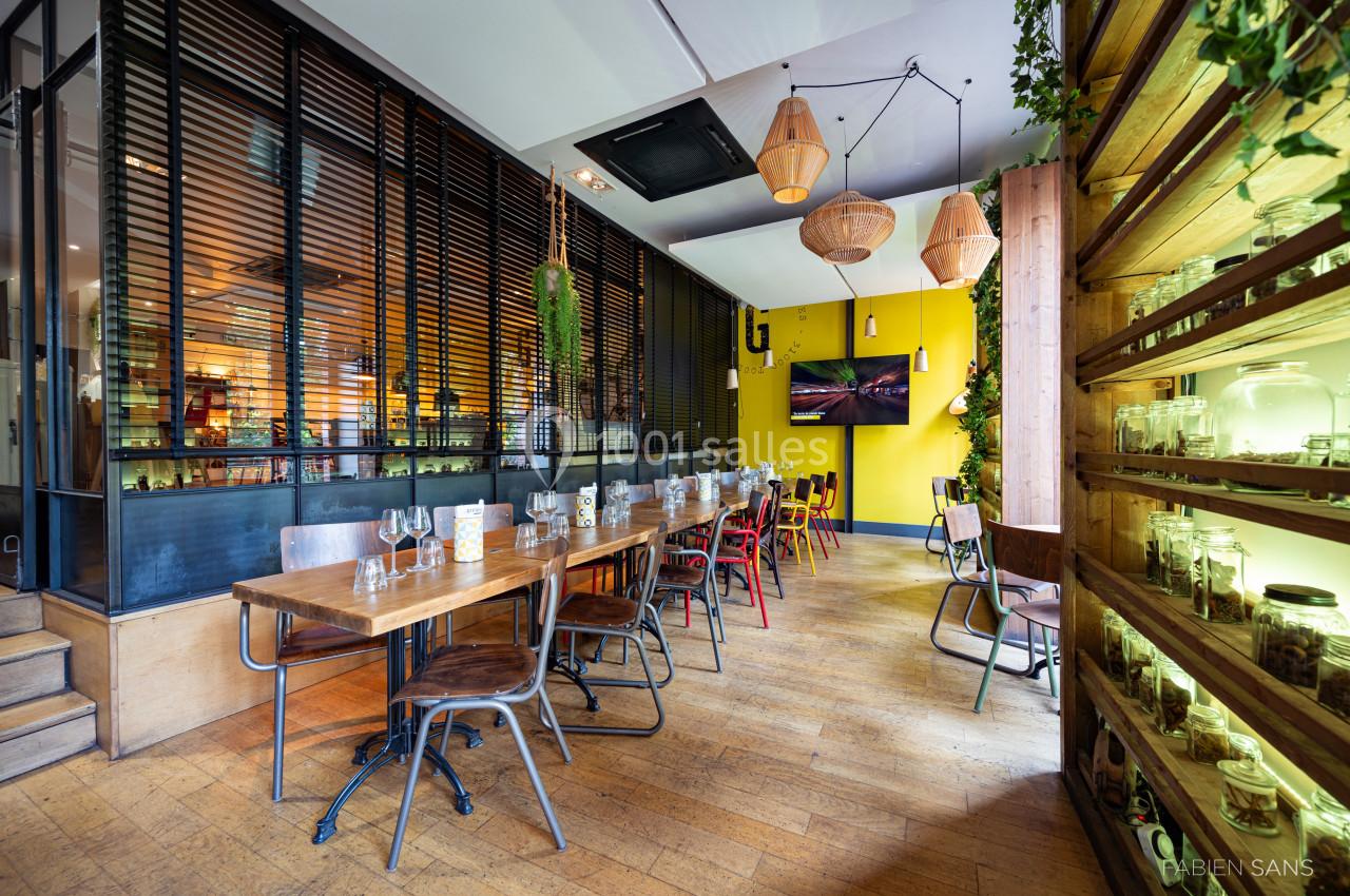 Salle de restaurant lumineuse avec tables en bois, chaises variées, murs jaunes et étagères décorées de bocaux.