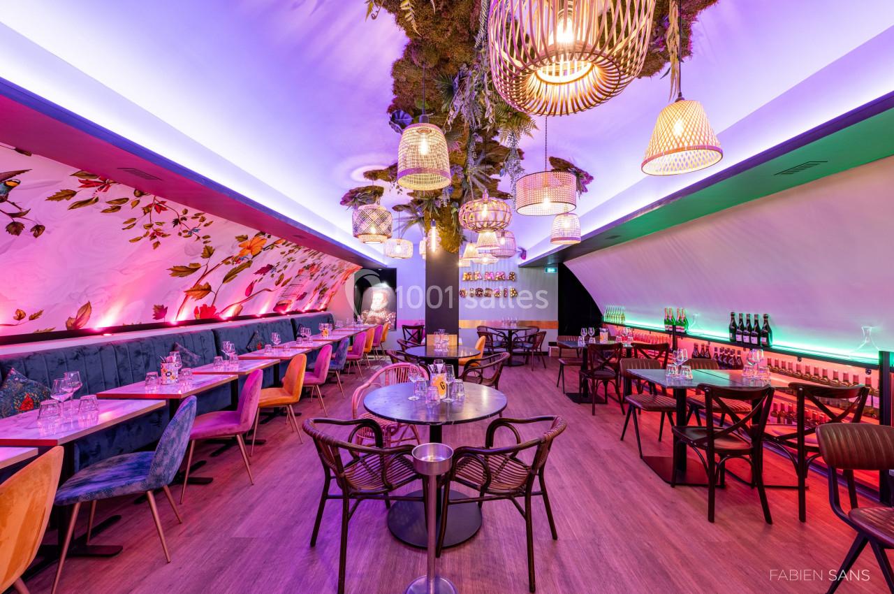 Salle de restaurant moderne avec tables, chaises, éclairage coloré et plafond décoré de végétation suspendue.