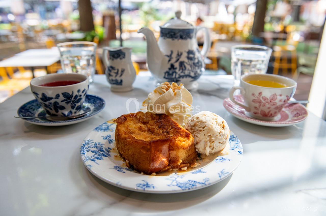 Assiette de pain perdu avec glace et chantilly, entourée de tasses de thé et d'une théière sur une table en terrasse.