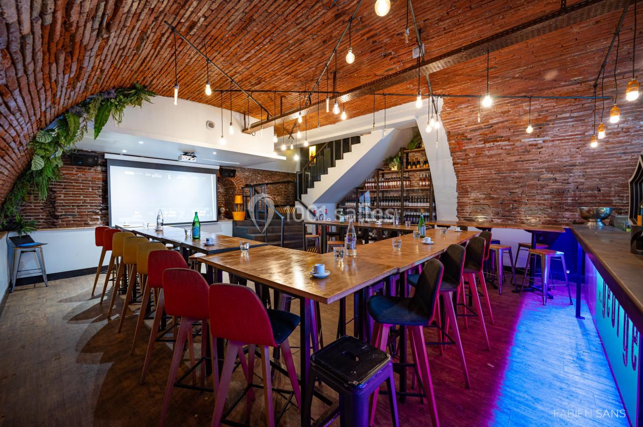 Salle voûtée en briques avec tables hautes, chaises colorées, éclairage suspendu et écran de projection.