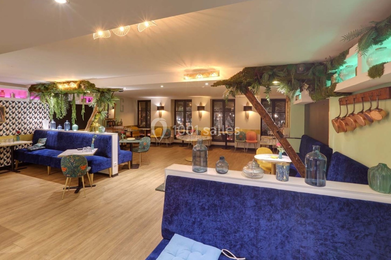 Salle lumineuse avec décoration végétale, banquettes bleues, tables colorées et accessoires en cuivre accrochés au mur.