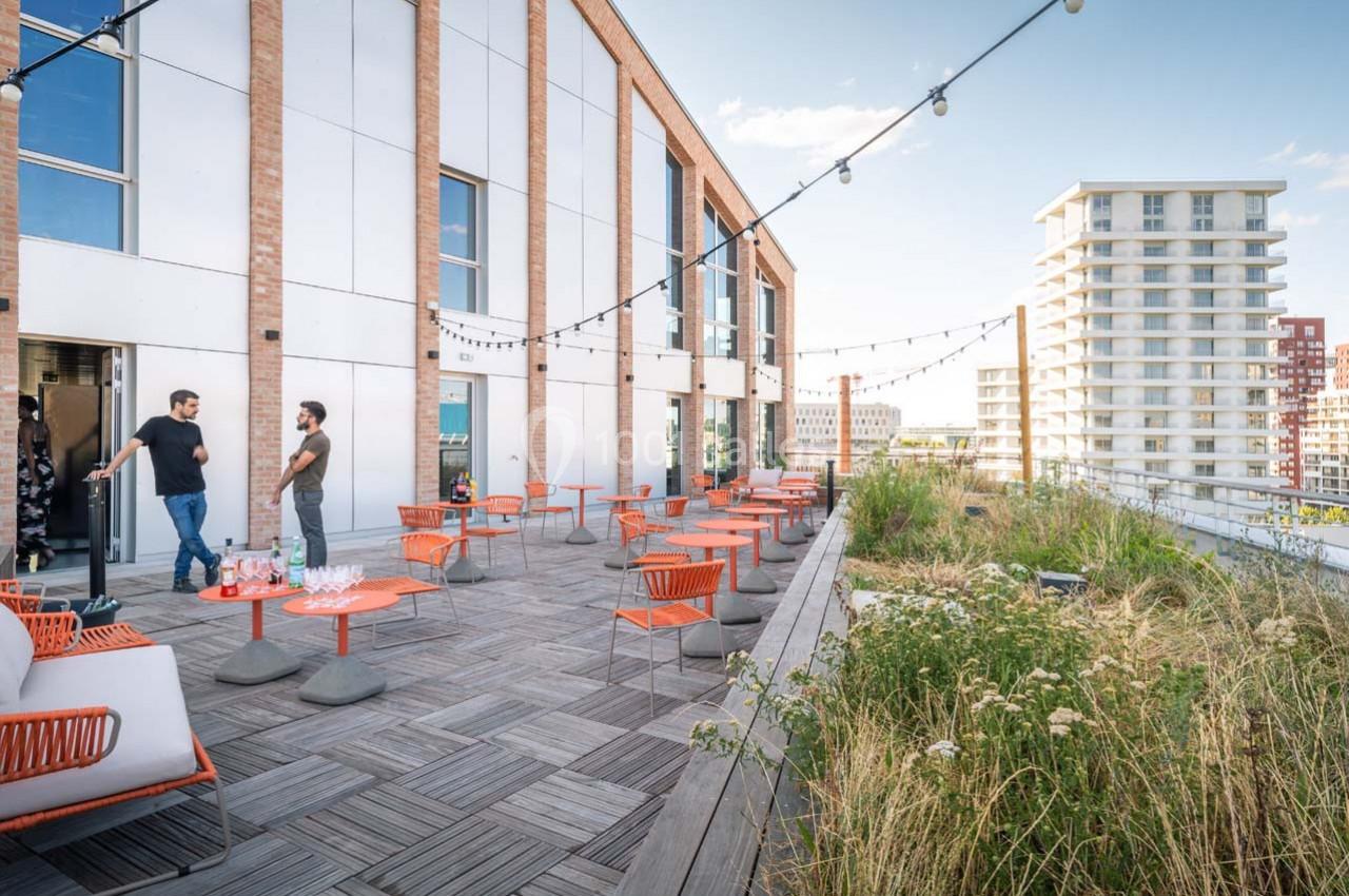 Location salle Ivry-sur-Seine (Val-de-Marne) - Walter Ivry #12 Terrasse en bois aménagée avec tables et chaises, entourée de végétation, donnant sur des immeubles modernes.