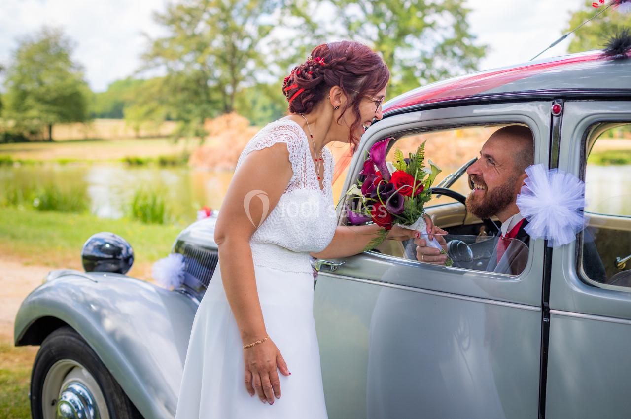 Une mariée souriante tend un bouquet à un marié assis dans une voiture ancienne près d'un étang.