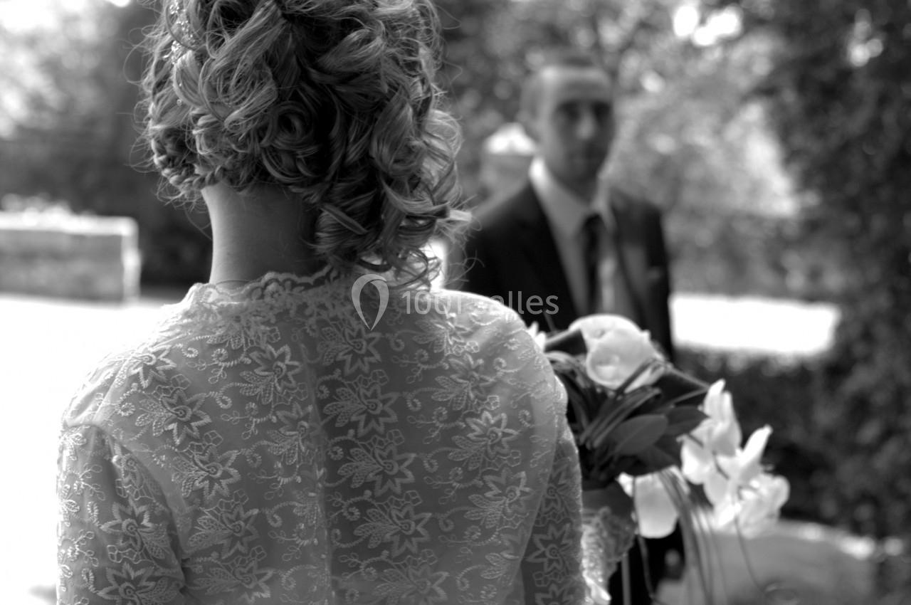 Une mariée de dos tenant un bouquet, face à un homme en costume dans un jardin en noir et blanc.