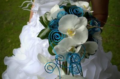 Femme en robe de mariée blanche tenant un bouquet de fleurs blanches et décorations bleues, en extérieur sur pelouse.