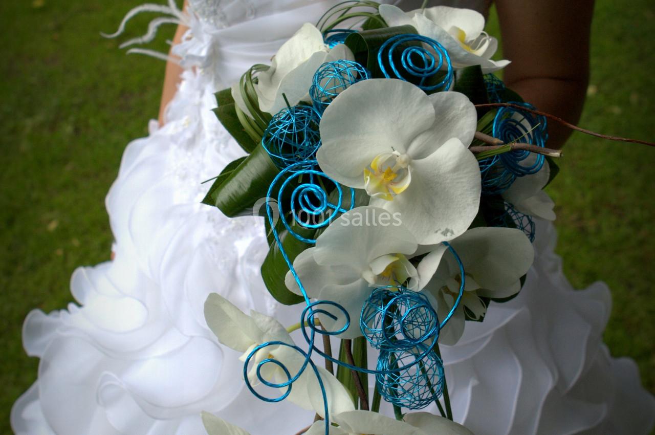 Femme en robe de mariée blanche tenant un bouquet de fleurs blanches et décorations bleues, en extérieur sur pelouse.