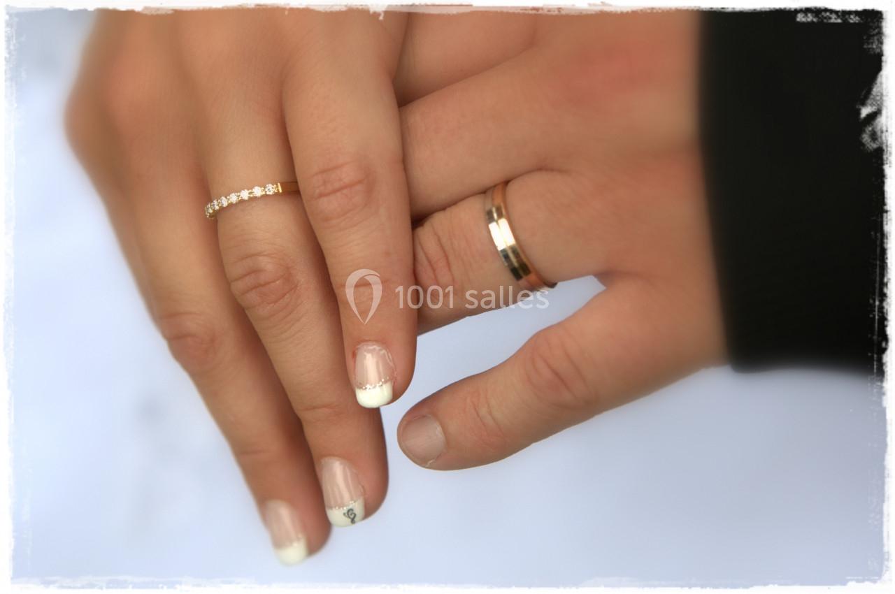 Deux mains jointes portant des alliances, l'une avec une manucure française, sur un fond flou.