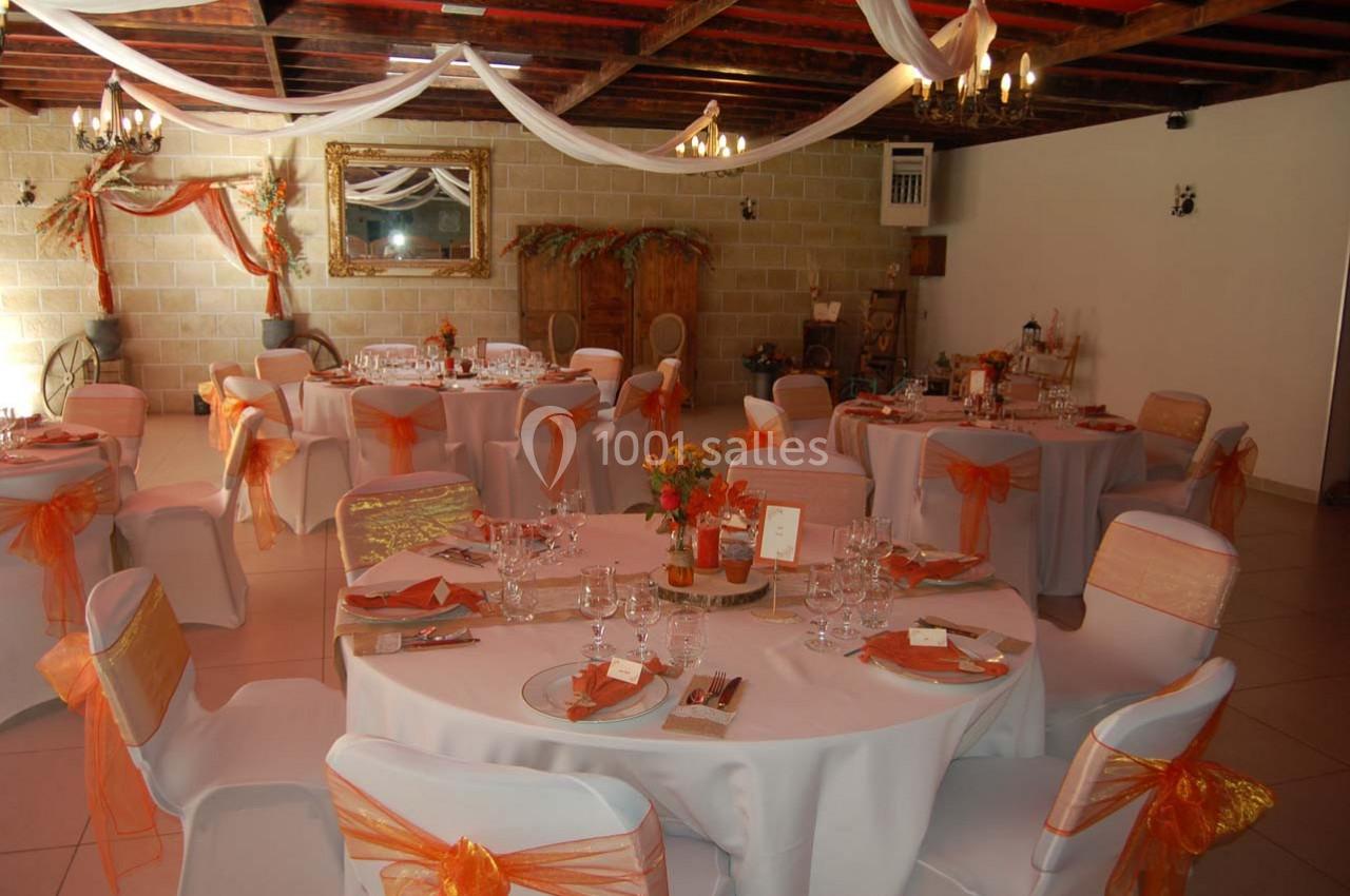 Salle de réception décorée avec des tables rondes, nappes blanches et chaises ornées de rubans orange.