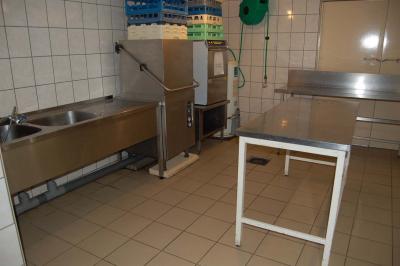 Cuisine professionnelle avec carrelage beige, plans de travail en inox, évier, réfrigérateur et éclairage fluorescent.