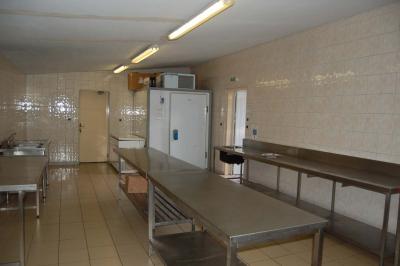 Cuisine professionnelle avec carrelage beige, plans de travail en inox, évier, réfrigérateur et éclairage fluorescent.