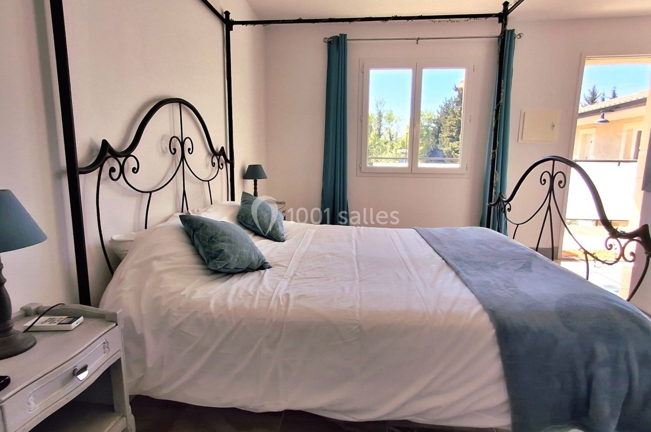 Chambre lumineuse avec lit double, linge blanc et bleu, deux tables de chevet et vue sur l'extérieur par une fenêtre.