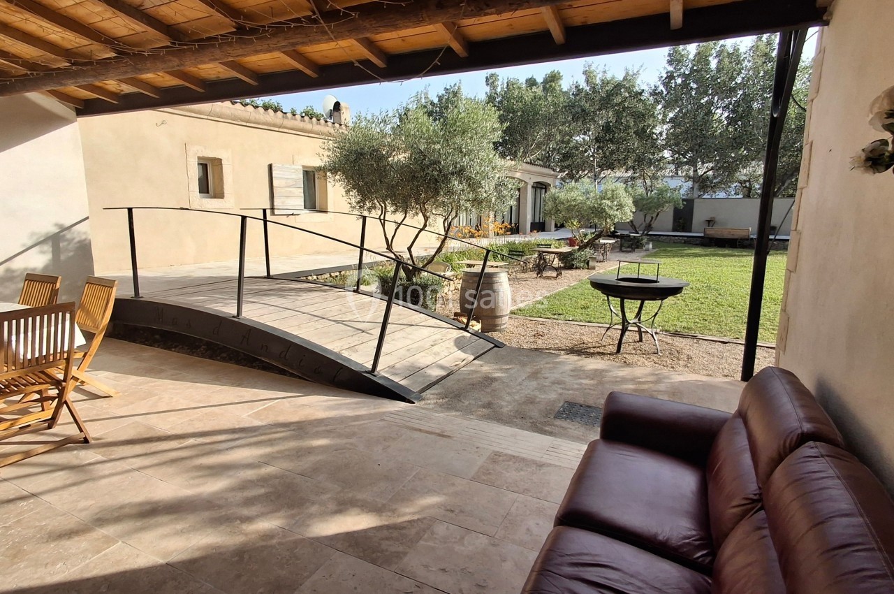 Terrasse avec salon de jardin, canapé, rampe en bois menant à un jardin arboré avec barbecue et oliviers.