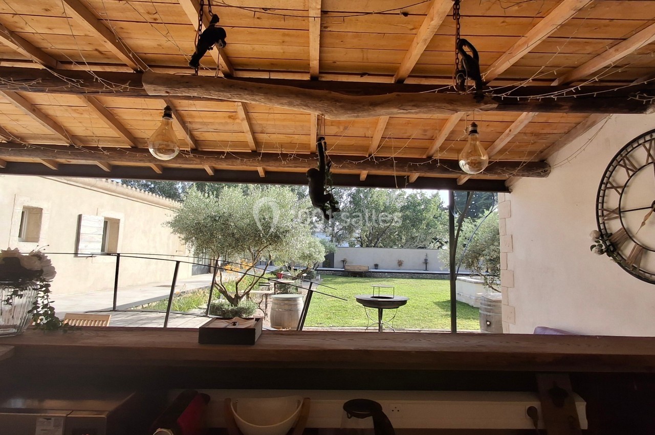 Vue depuis un bar en bois sous une pergola, donnant sur un jardin avec olivier, barbecue et horloge murale.