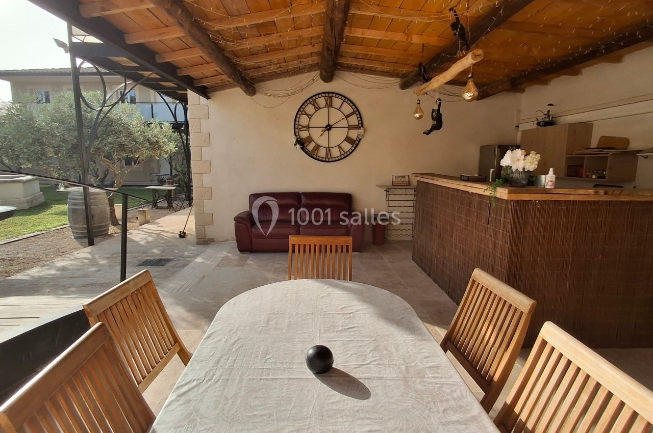 Espace extérieur couvert avec table, chaises en bois, canapé, bar en carrelage et horloge murale décorative.