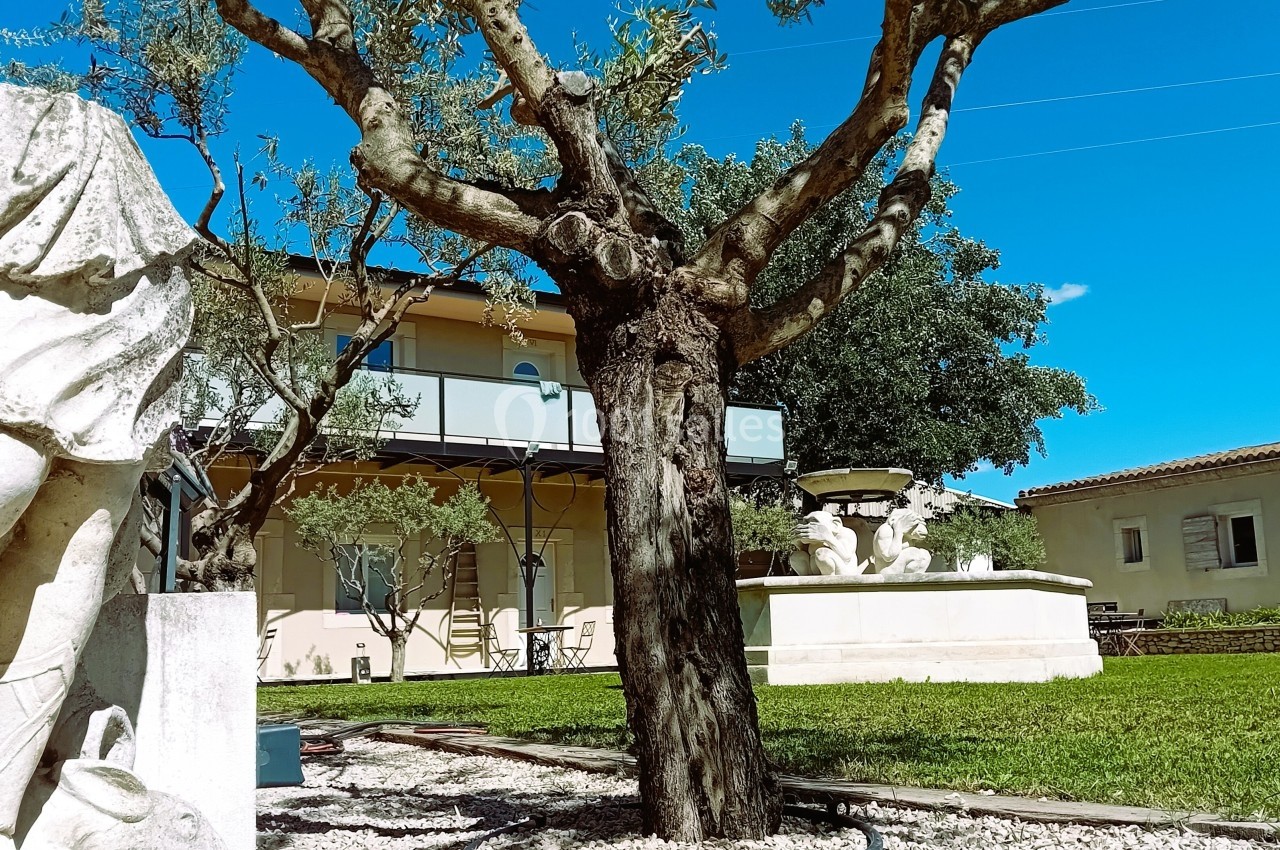 Statue en pierre, arbre et fontaine dans un jardin avec pelouse, devant un bâtiment beige sous un ciel bleu.