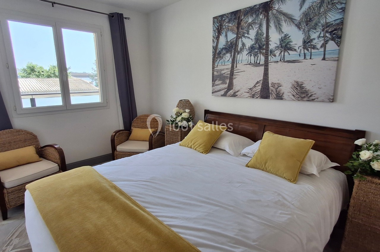 Chambre lumineuse avec un lit double, coussins jaunes, fauteuils en rotin et tableau représentant une plage tropicale.