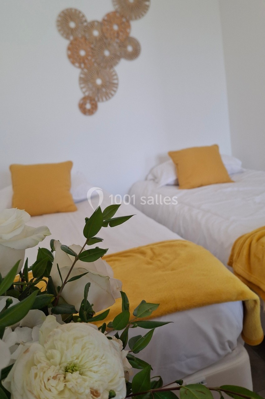 Deux lits simples avec linge blanc et coussins jaunes, décorés de fleurs blanches au premier plan et d'un mur orné.
