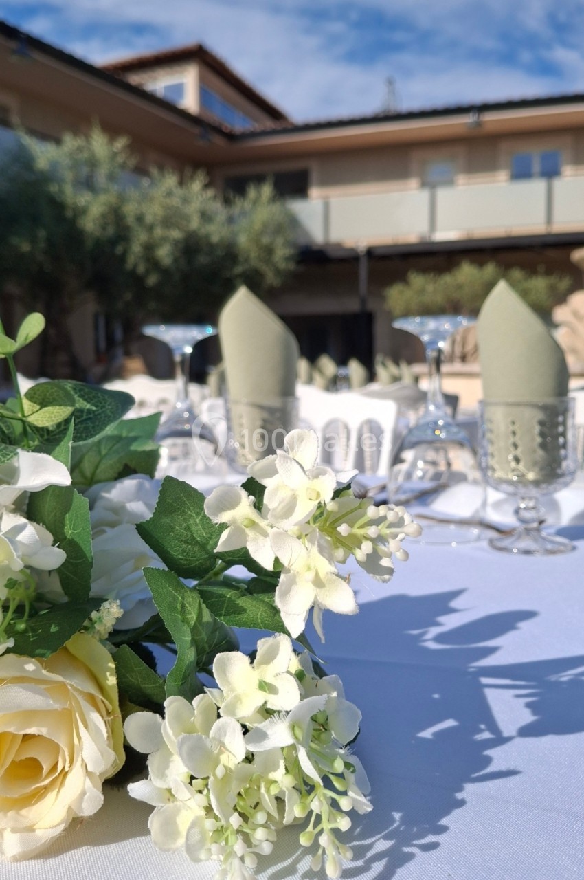 Table dressée en extérieur avec nappes blanches, fleurs blanches et jaunes, verres et serviettes pliées, devant un bâtiment.