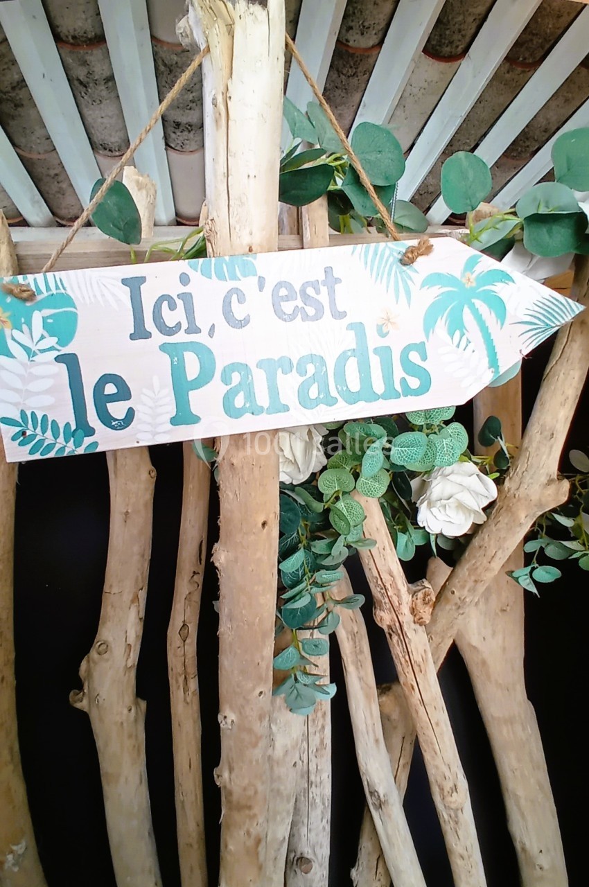 Pancarte en bois suspendue avec l'inscription ’Ici, c'est le Paradis’, entourée de branches et de feuillage.