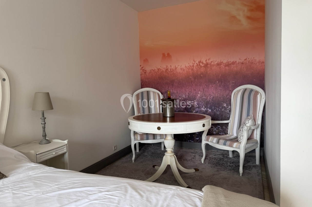 Chambre avec un lit, une table ronde, deux fauteuils rayés et un mur décoré d'une photo de paysage au coucher de soleil.