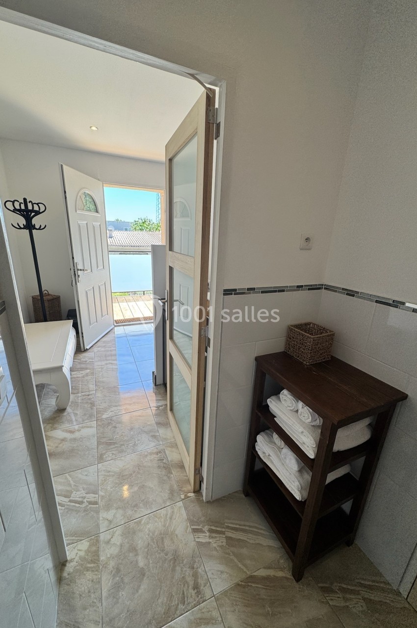 Salle de bain lumineuse avec étagère en bois, serviettes pliées et porte ouverte sur une terrasse ensoleillée.