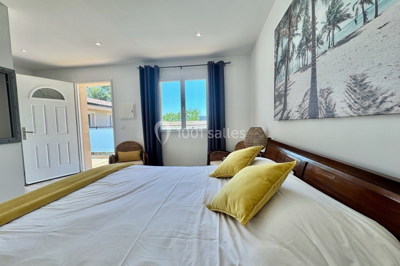 Chambre lumineuse avec lit double, coussins jaunes, fauteuils en osier et tableau représentant une plage tropicale.