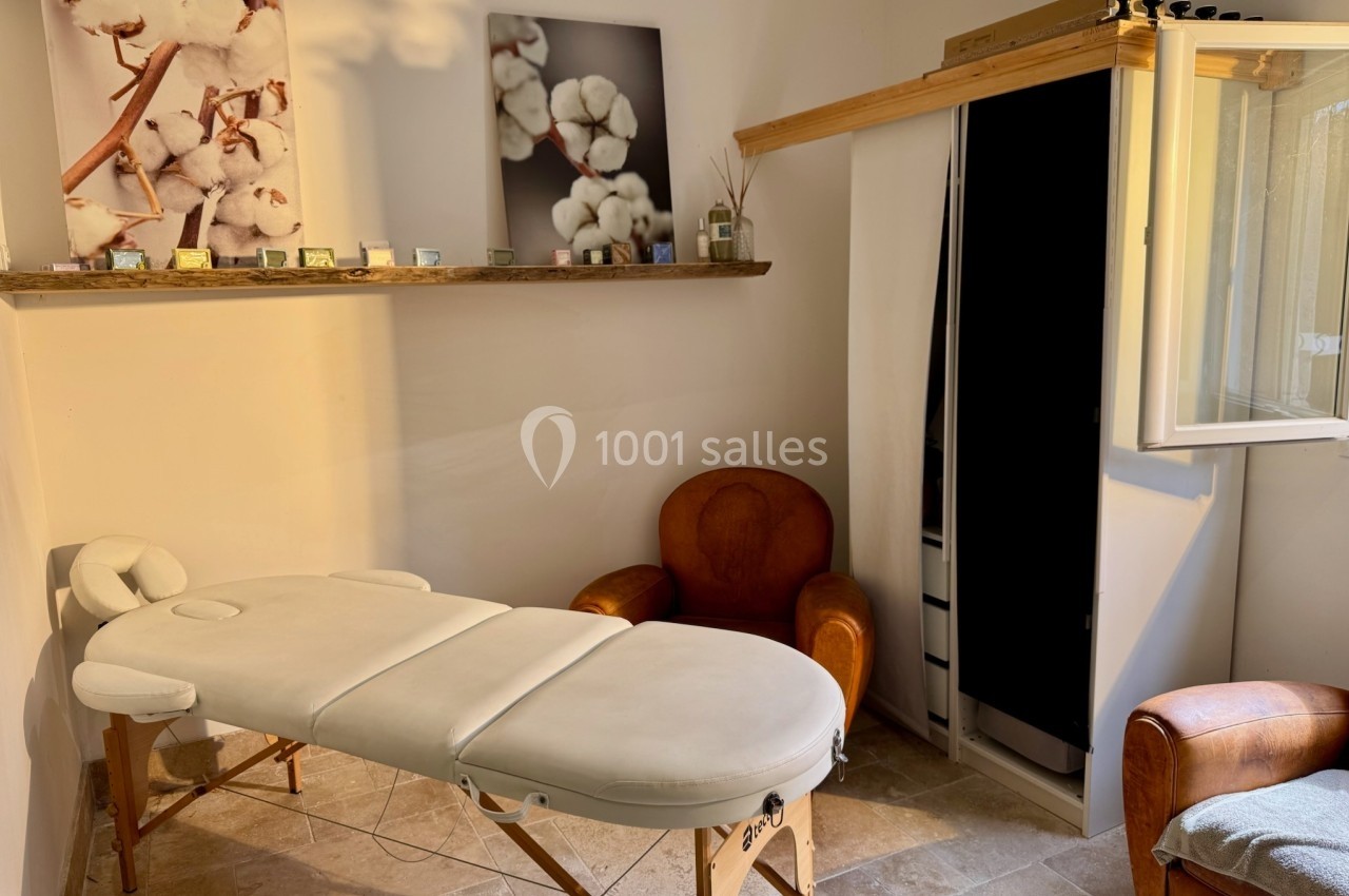 Salle lumineuse avec table de massage, fauteuils en cuir, étagère décorée et cadres représentant des fleurs de coton.