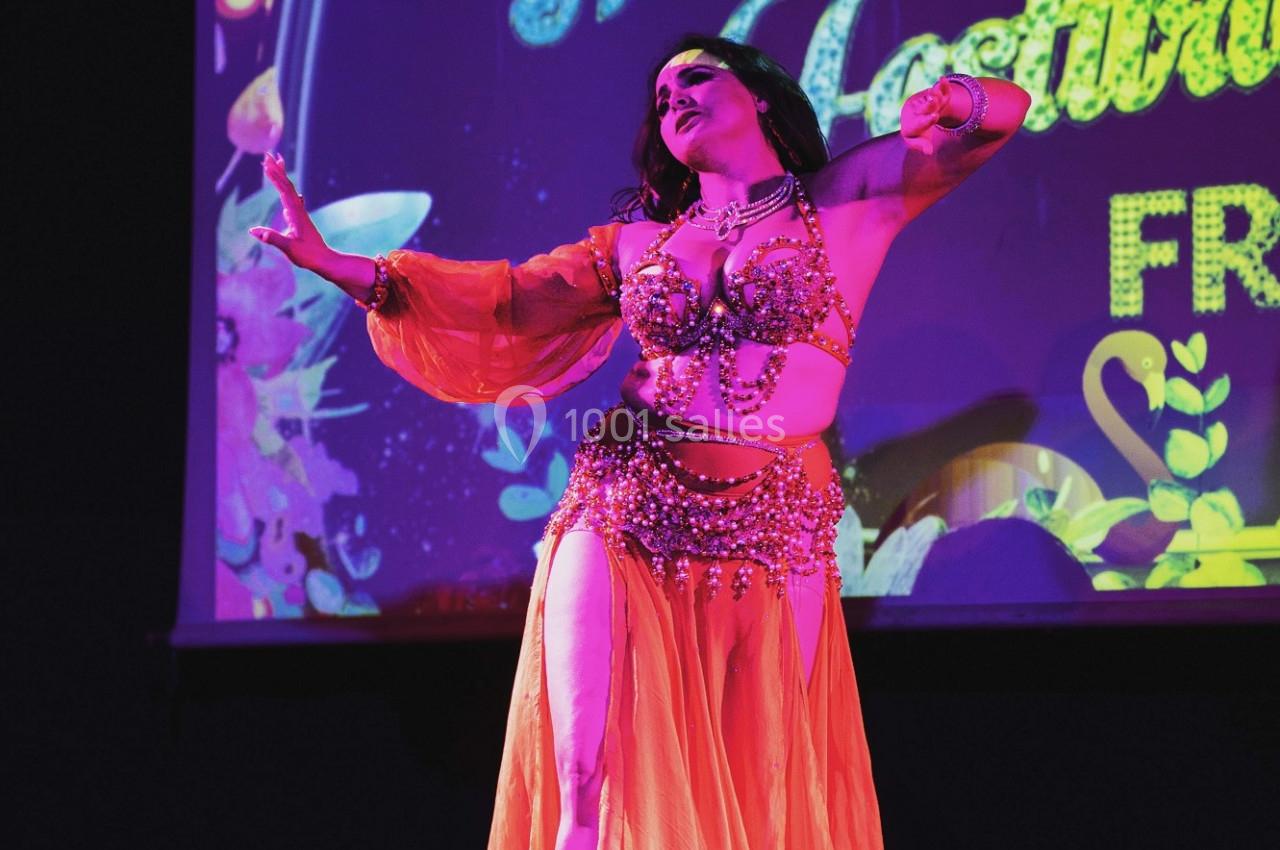 Danseuse en tenue orientale orange exécutant une performance sur une scène éclairée avec un fond coloré.
