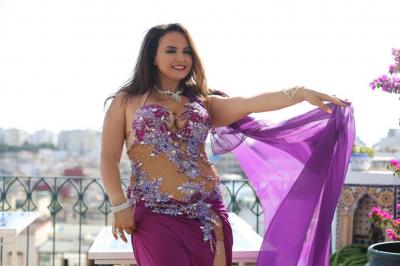 Femme en tenue de danse orientale violette, posant sur une terrasse ensoleillée décorée de plantes et de mobilier extérieur.